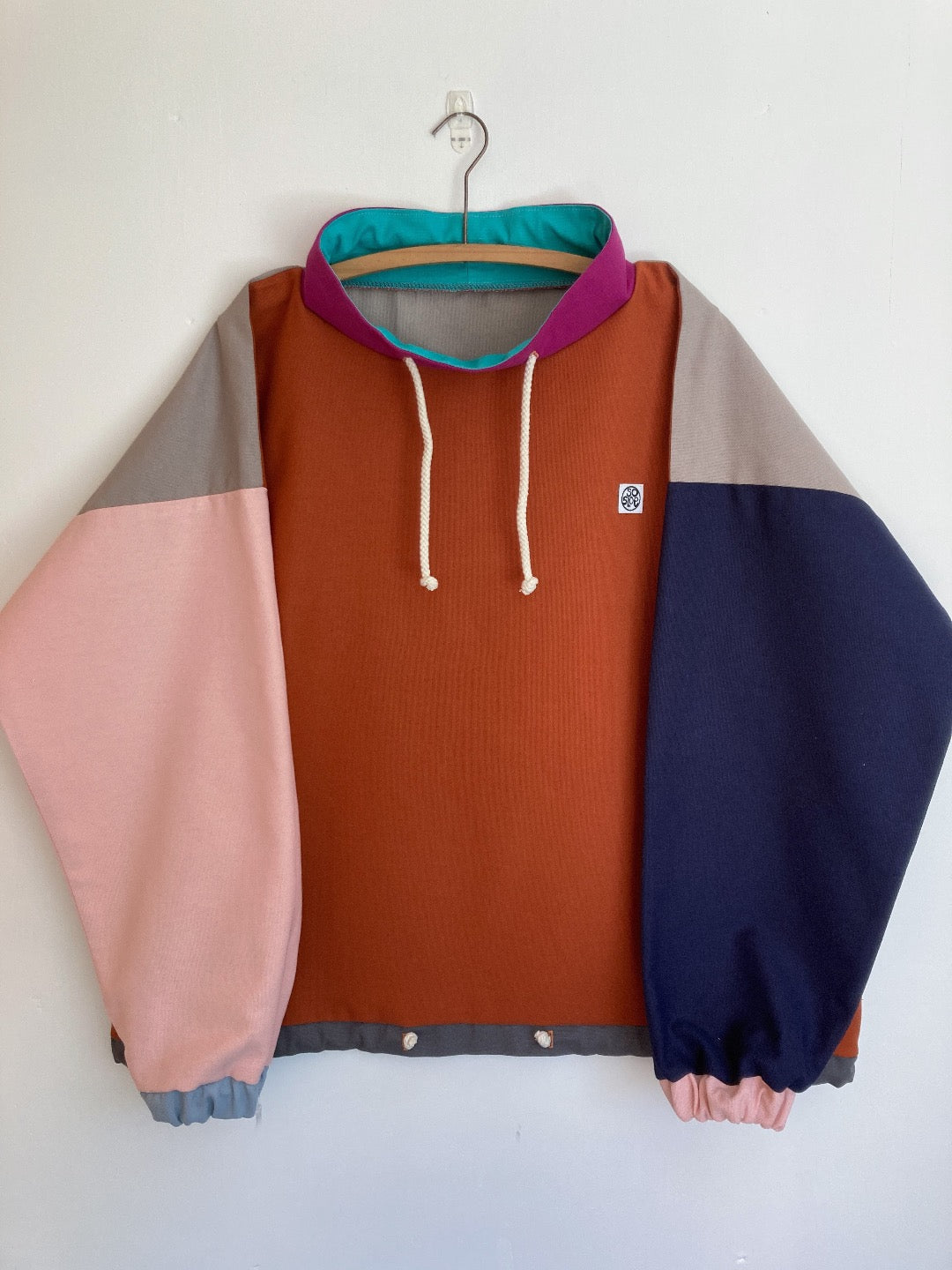 Cotton pullover in orange with one pale pink sleeve and one navy blue sleeve. Grey back panel and fuchsia pink collar with drawstring. 